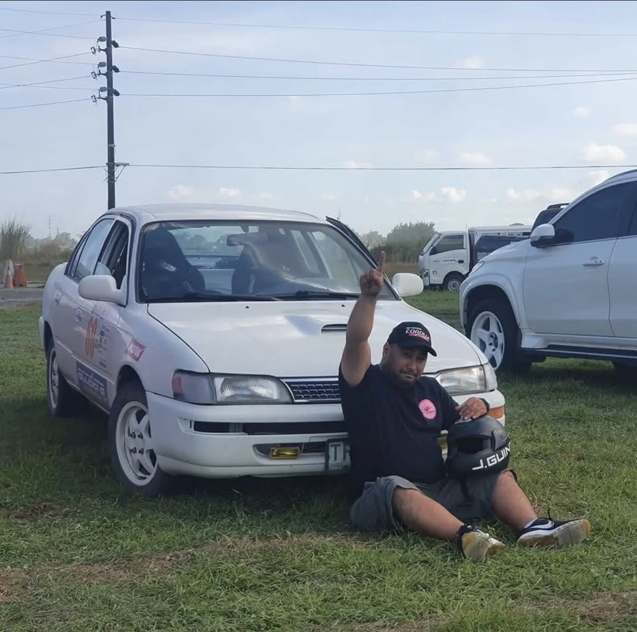 Jonathan Guintu sitting beside his white Toyota Corolla race car, one finger raised